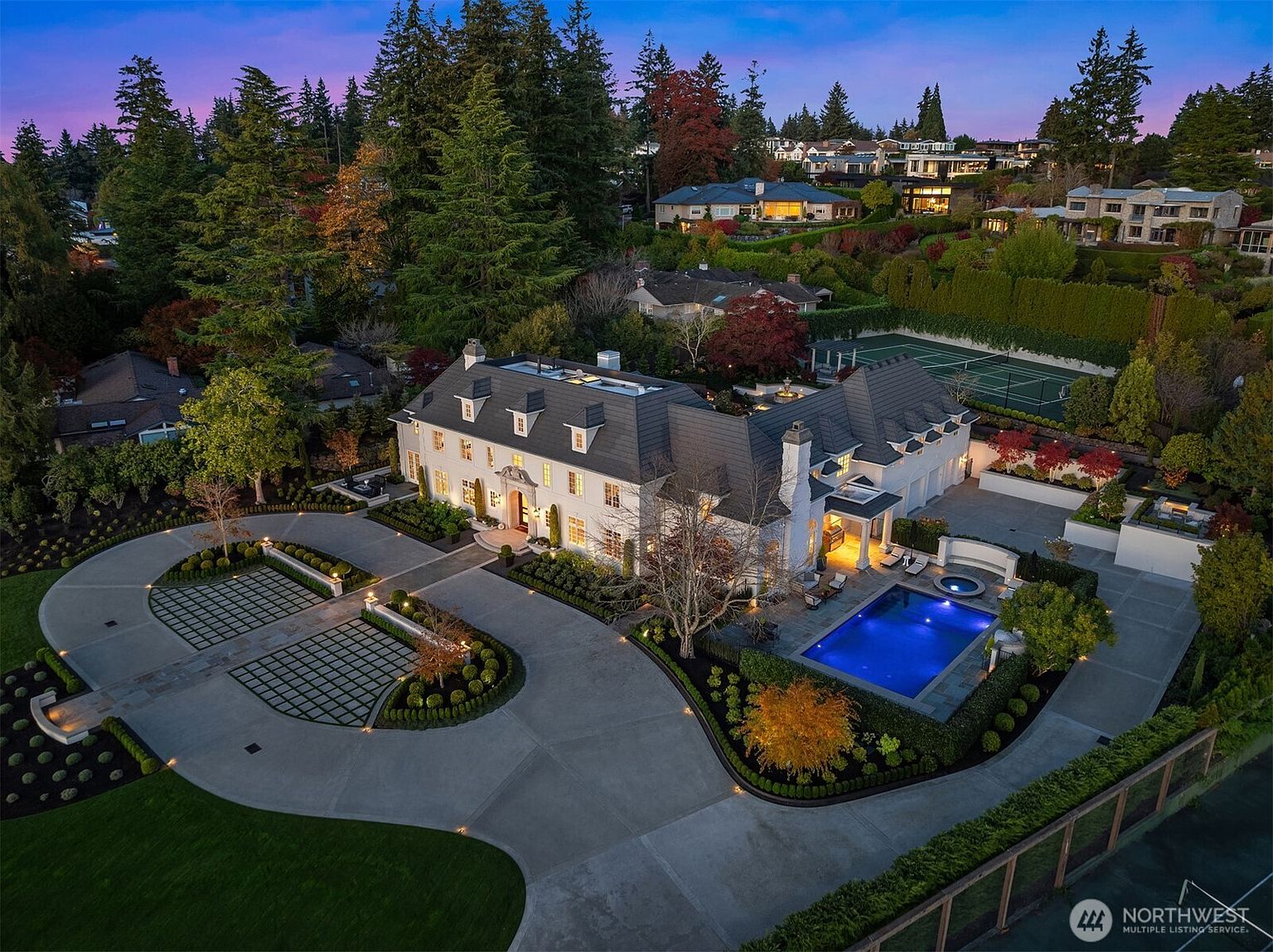 This stunning aerial perspective captures a grand, multi-story estate at twilight, showcasing its sophisticated architectural design and expansive grounds. The property features a meticulously landscaped circular driveway, a vibrant blue swimming pool with an integrated spa, and a private tennis court nestled among lush, mature trees. The warm glow from the home's interior lighting contrasts beautifully with the deep blue evening sky, creating an inviting and luxurious atmosphere.