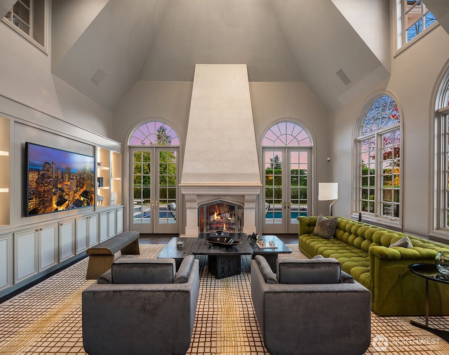 This elegant, high-ceilinged living room features a grand stone fireplace as its focal point, flanked by symmetrical arched glass doors that open to a pool area. The space is anchored by a vibrant green tufted sofa and two plush grey armchairs, all set upon a geometric patterned rug. Built-in cabinetry with an integrated television adds a modern touch to the sophisticated, airy design.