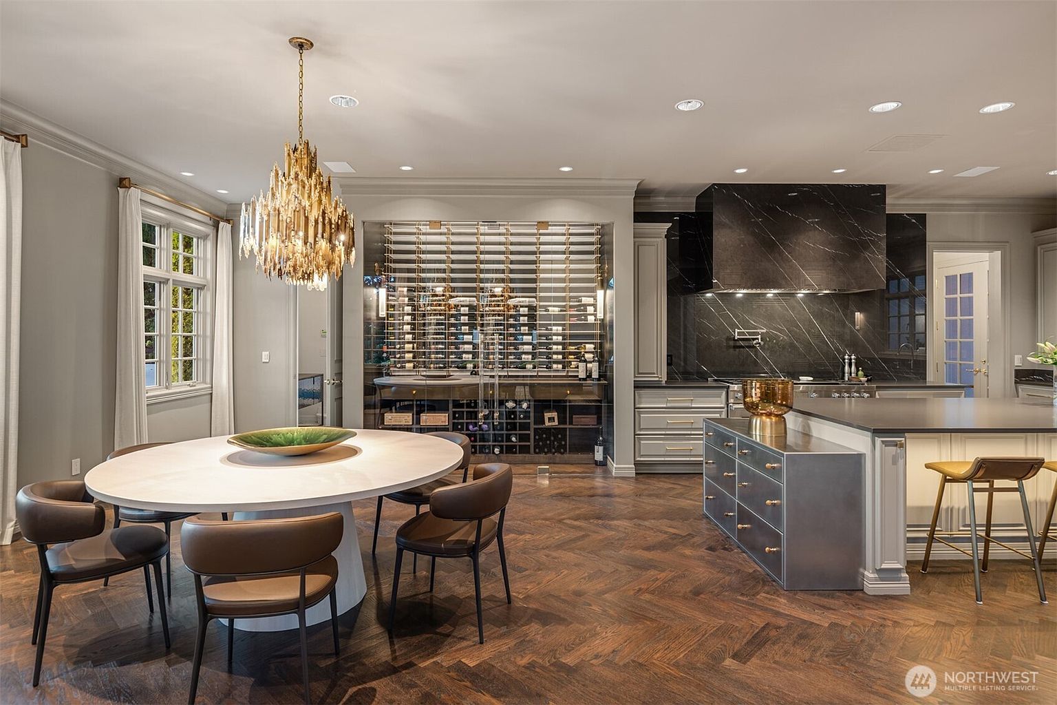 This sophisticated, open-concept kitchen features a large central island with a metallic finish, a striking black marble backsplash, and a custom built-in wine cellar. A round dining table with modern chairs sits under a dramatic gold chandelier, all set against rich, dark herringbone hardwood floors. The space exudes a luxurious, high-end aesthetic with a seamless blend of contemporary design and elegant textures.