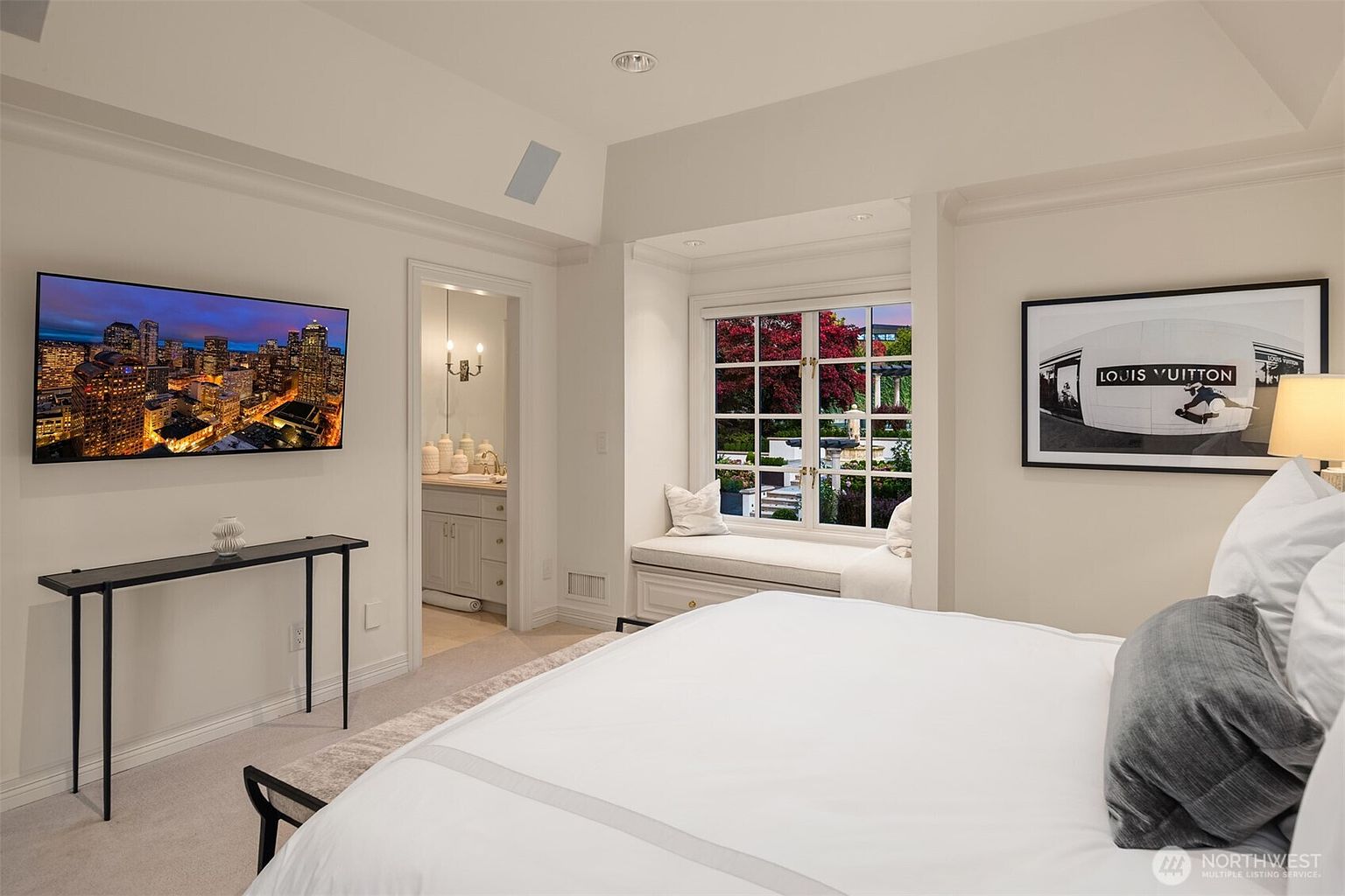 This elegant primary bedroom features a neutral color palette, a plush bed with crisp white linens, and a cozy window seat nestled within a recessed alcove. A sleek, minimalist console table sits beneath a large wall-mounted television, while a framed Louis Vuitton art piece adds a touch of sophisticated branding to the wall. The room offers a seamless transition into an adjoining bathroom, creating a bright, airy, and luxurious atmosphere.