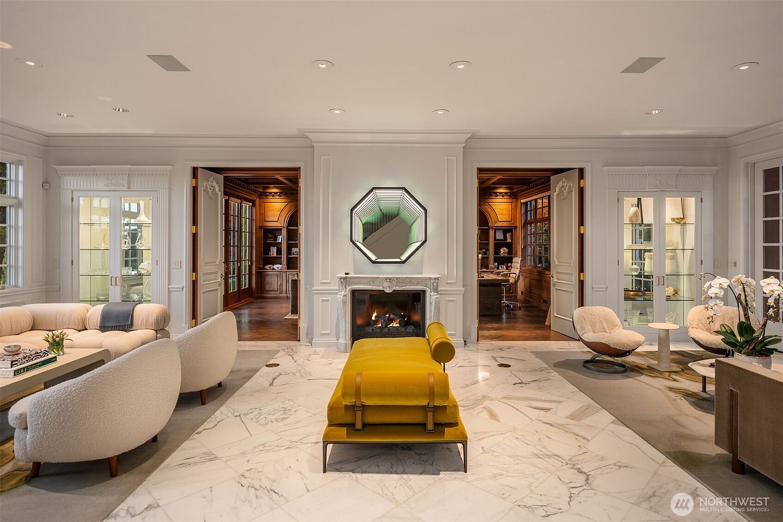This elegant, symmetrical living room features a striking marble floor and a central fireplace adorned with a unique geometric infinity mirror. The space is flanked by two sets of double doors leading into wood-paneled studies, creating a sophisticated and balanced atmosphere. A vibrant yellow daybed serves as a bold focal point, complemented by plush, curved seating and high-end decor that exudes luxury and refined taste.