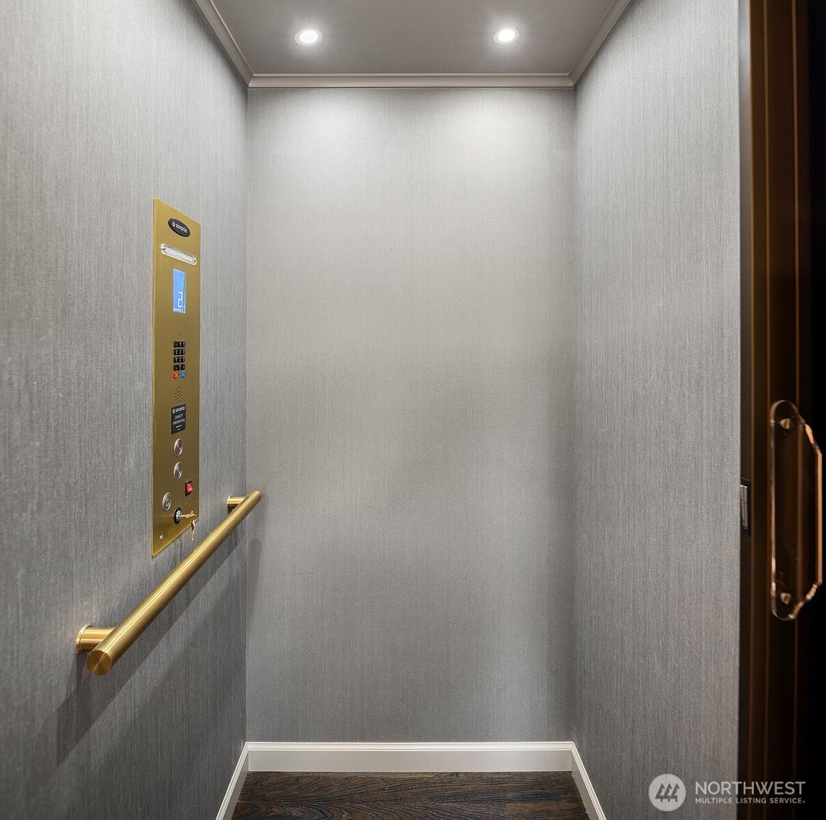 This image shows the interior of a private residential elevator, featuring a sleek, modern design with textured grey wallpaper and a polished brass control panel. A matching brass handrail runs along the side wall, and the space is illuminated by two recessed ceiling lights, creating a sophisticated and minimalist aesthetic. The perspective is a straight-on view looking into the elevator car, highlighting its clean lines and high-end finishes.