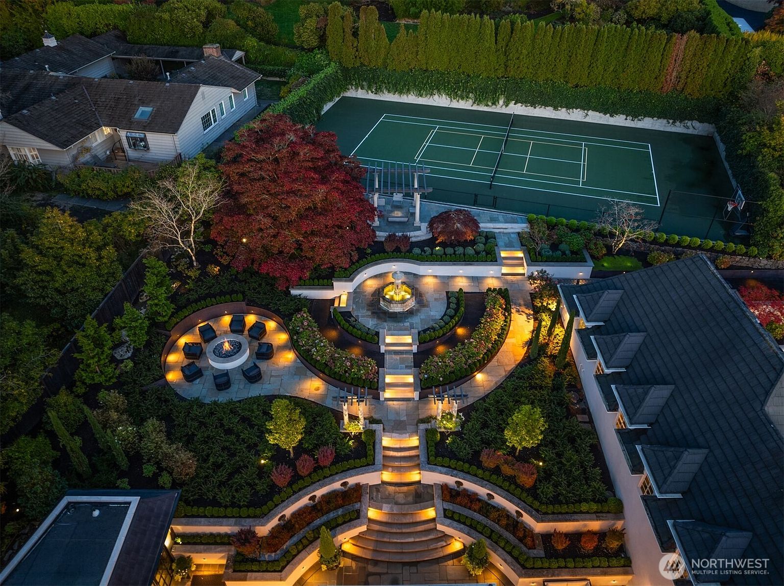 This stunning aerial view captures a meticulously landscaped backyard at twilight, featuring a multi-tiered stone patio, a circular fire pit lounge, and a central fountain. The property includes a private tennis court bordered by tall hedges, creating a secluded and luxurious outdoor retreat. The warm glow of integrated landscape lighting highlights the architectural pathways and lush greenery, emphasizing the home's sophisticated design.