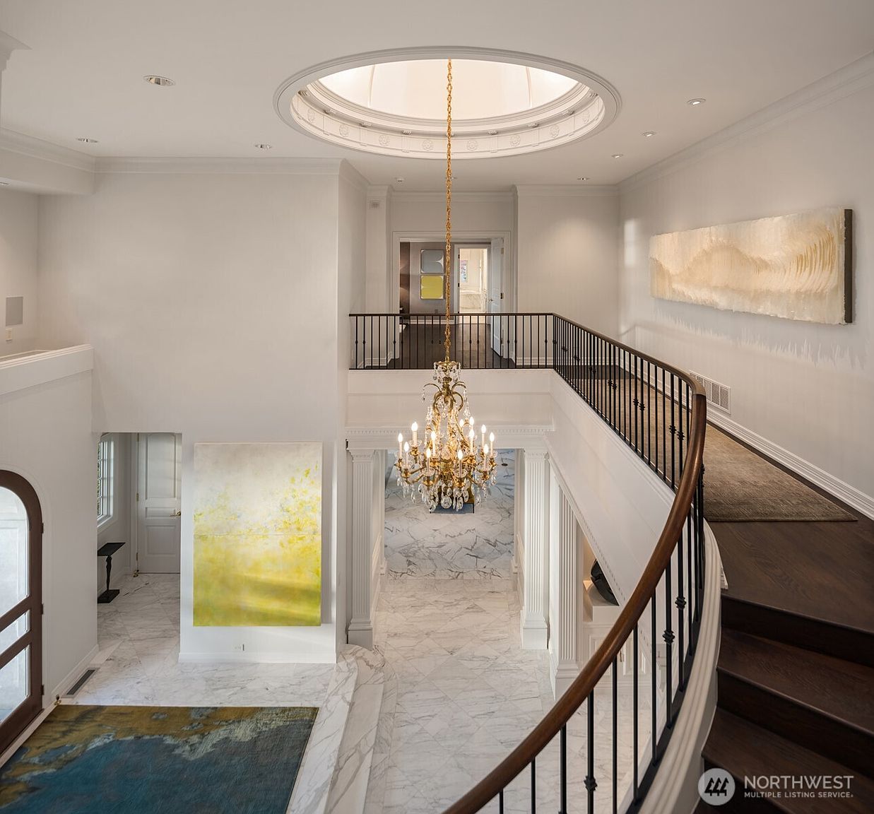 This grand foyer features a sweeping curved staircase with dark wood treads and a wrought-iron railing, overlooking a luxurious marble-floored entryway. A magnificent crystal chandelier hangs from a dramatic circular skylight, casting light over the open-concept space and large, vibrant abstract art pieces. The perspective is from the upper landing, emphasizing the architectural elegance and the airy, high-ceilinged design of the home.