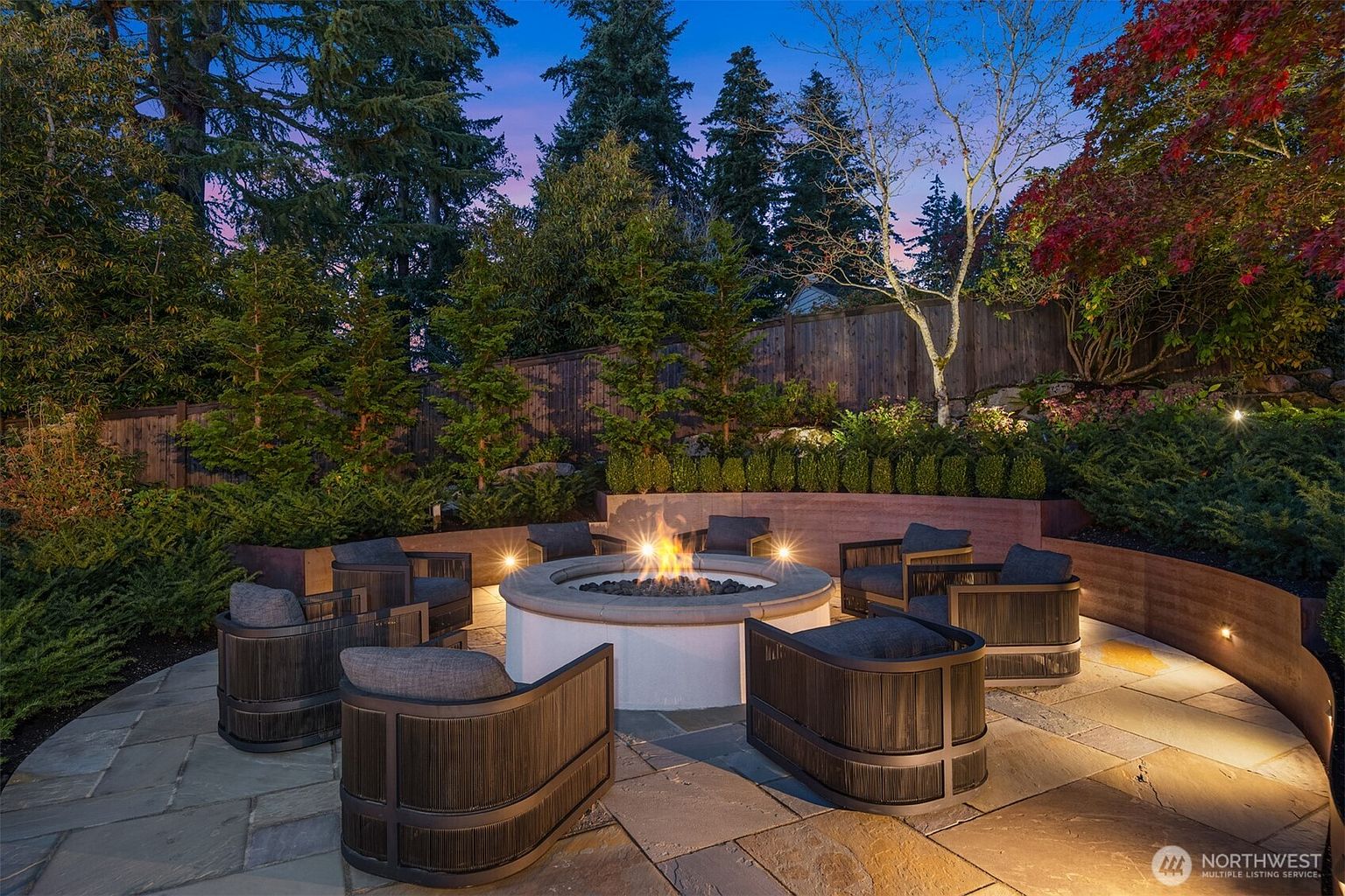 This inviting outdoor patio features a circular stone fire pit as the centerpiece, surrounded by six modern, curved-back lounge chairs with plush cushions. The space is set against a tiered landscape with a retaining wall, lush greenery, and mature trees, all illuminated by warm accent lighting that creates a cozy, sophisticated evening atmosphere. The flagstone paving and thoughtful landscaping make this an ideal area for outdoor entertaining and relaxation.