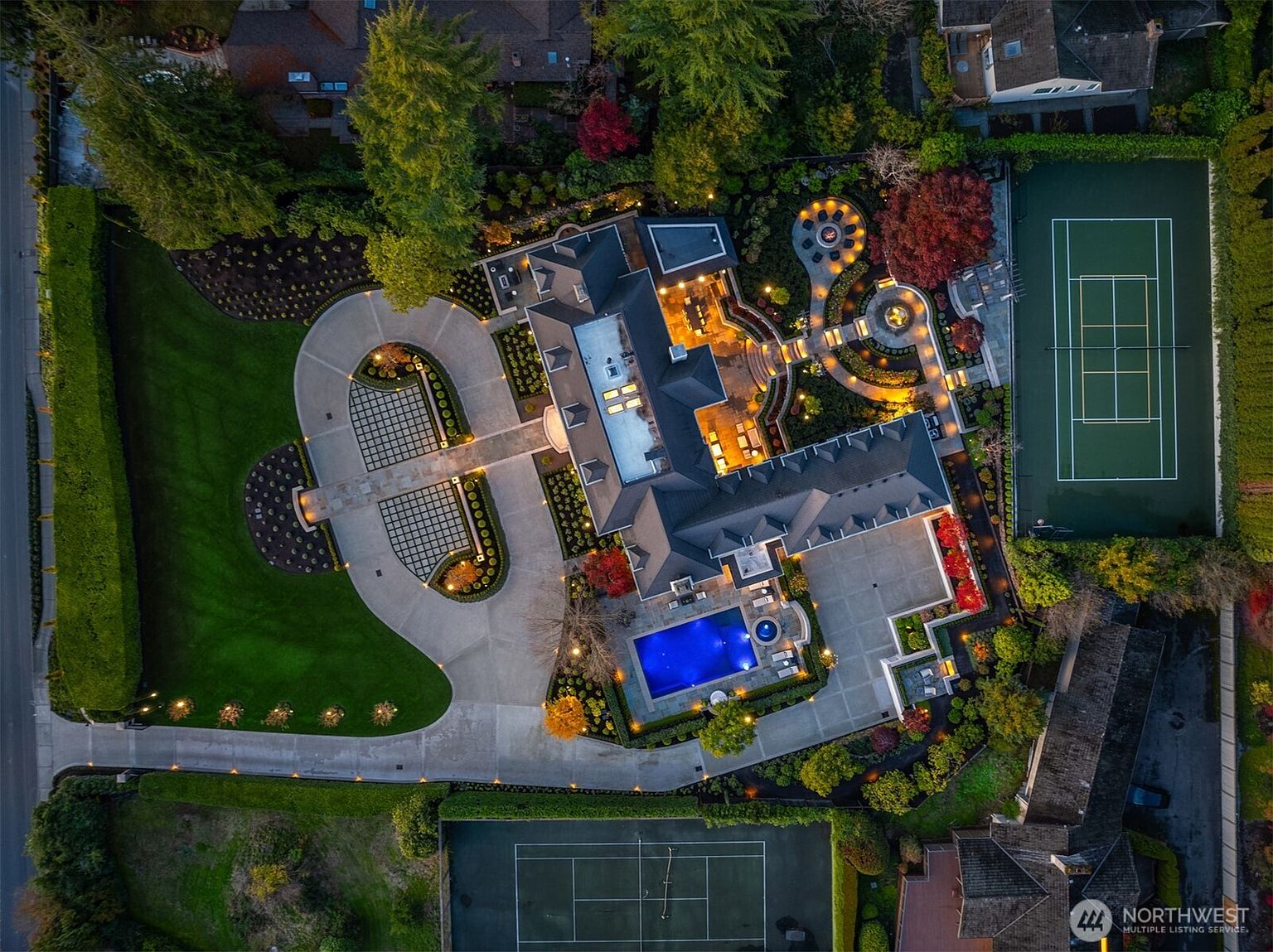 This stunning aerial view captures a luxurious estate at twilight, showcasing a sprawling residence with a sophisticated architectural design. The property features a vibrant blue swimming pool, two private tennis courts, and meticulously landscaped gardens illuminated by warm, ambient lighting. The expansive driveway and lush greenery create an impression of exclusivity and high-end living.