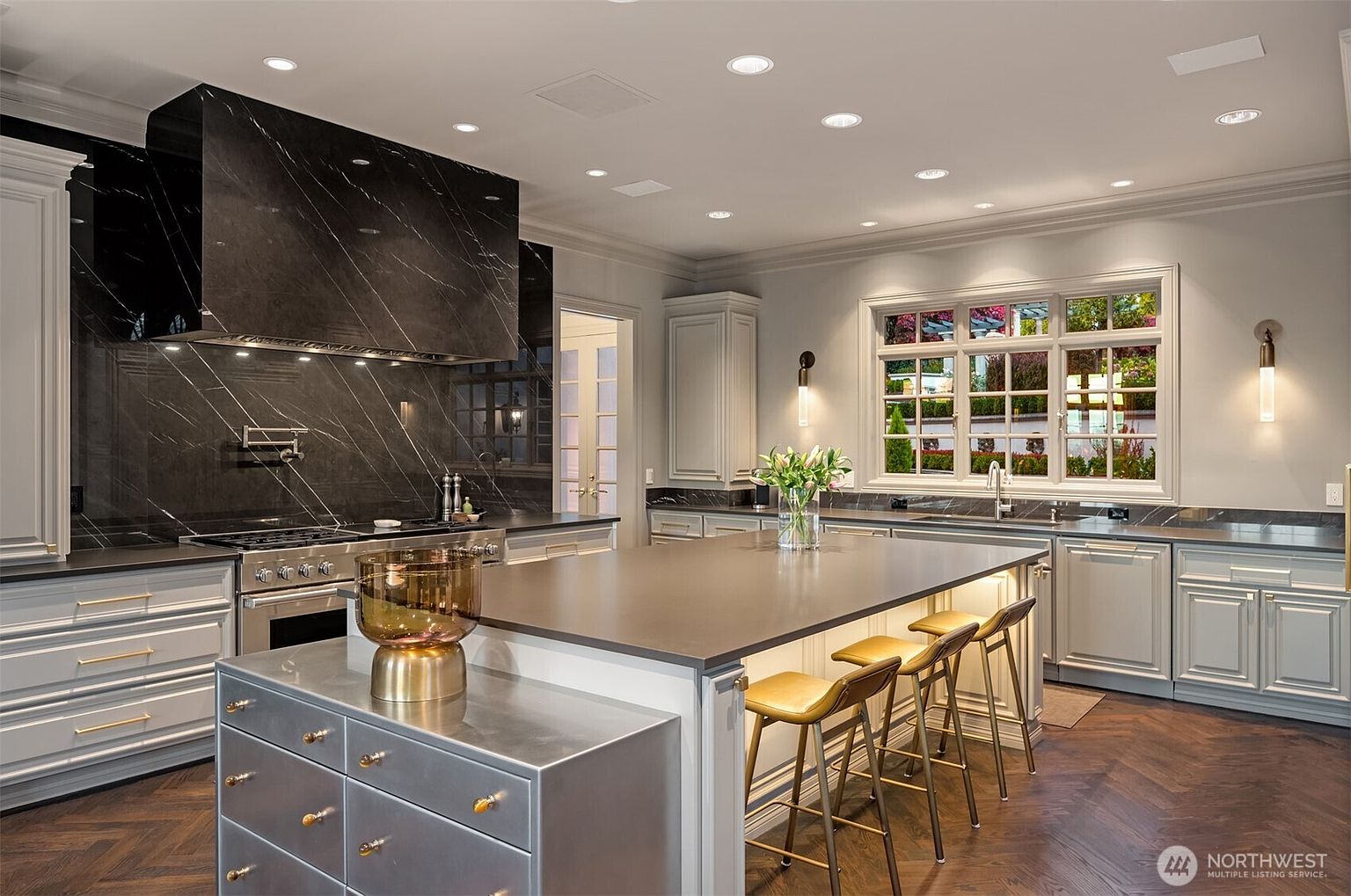This sophisticated, high-end kitchen features a striking black marble backsplash and range hood that contrast beautifully with the light grey cabinetry and dark herringbone wood flooring. A large central island with a matte countertop and metallic accents serves as the focal point, complemented by three stylish bar stools. The space is brightened by recessed lighting and a large window, creating an elegant and functional culinary environment.