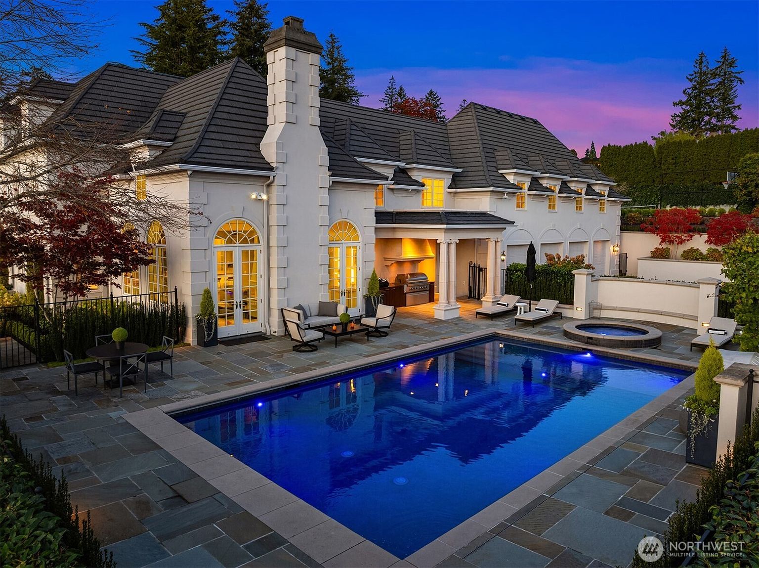 This stunning rear exterior view showcases a luxurious estate featuring a large, illuminated swimming pool and an integrated hot tub set within a stone-paved patio. The home displays elegant architectural details, including arched glass doors, a prominent stone chimney, and a covered outdoor kitchen area, all set against a vibrant twilight sky. The scene exudes a sophisticated and tranquil atmosphere, perfect for high-end outdoor entertaining.
