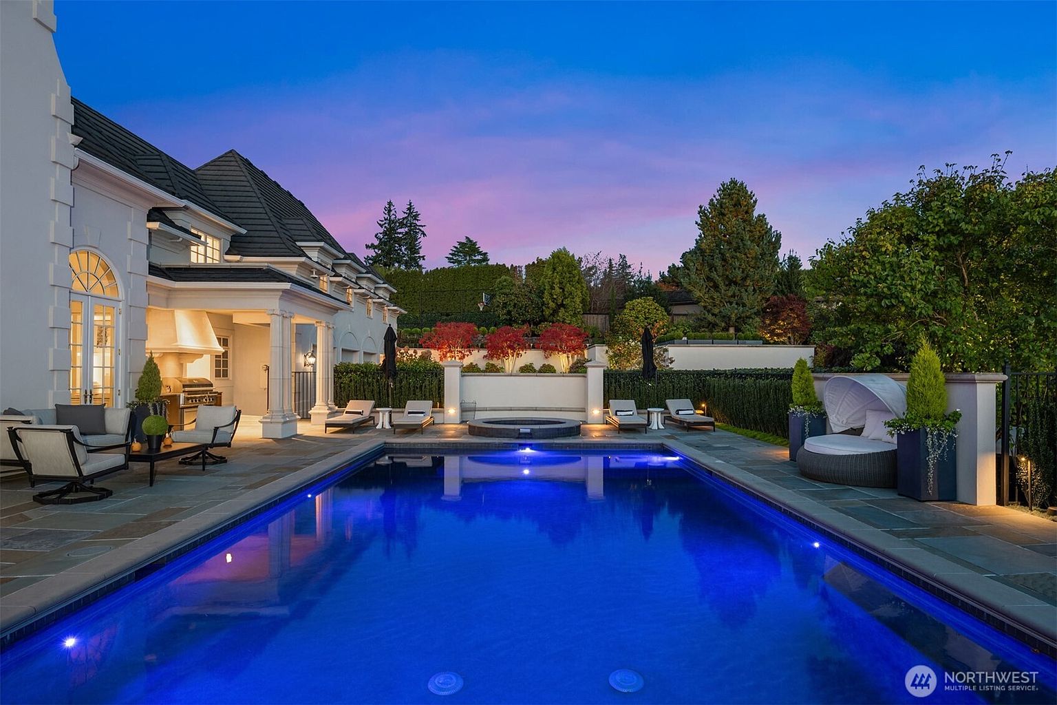 This stunning backyard oasis features a vibrant, illuminated rectangular swimming pool as the centerpiece, complemented by a built-in spa and a spacious stone patio. The area is elegantly furnished with lounge chairs and a cozy daybed, all set against the backdrop of a sophisticated home exterior and lush, manicured landscaping under a dramatic twilight sky. The scene exudes luxury and tranquility, perfect for high-end outdoor entertaining.