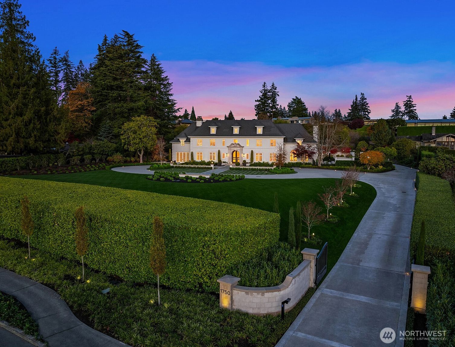 This stunning aerial perspective captures a grand, white colonial-style estate at twilight, set against a vibrant sunset sky. The property features a meticulously manicured lawn, a circular driveway, and a tall, perfectly trimmed hedge that frames the front entrance. The elegant architecture, combined with the warm glow of the interior lights and landscape lighting, creates a sophisticated and inviting atmosphere.