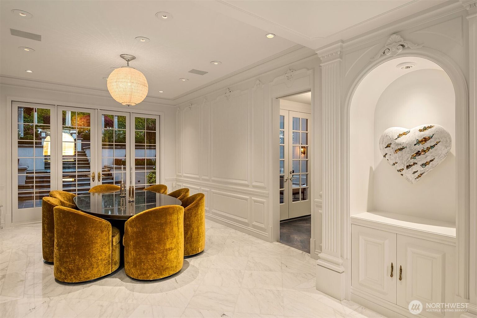 This elegant dining room features a sophisticated, monochromatic white palette with intricate wall molding and polished marble flooring. A striking round glass-top table is surrounded by plush, mustard-yellow velvet chairs, while a large, textured heart-shaped art piece serves as a focal point within a recessed arched niche. The room is bathed in soft, warm light from a contemporary globe pendant fixture, creating a luxurious and inviting atmosphere.