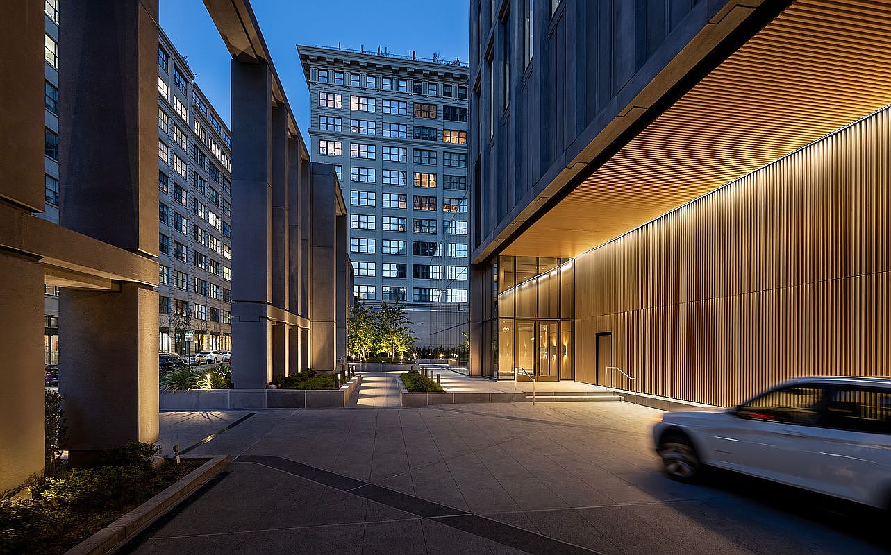 This sophisticated urban entryway features a dramatic, modern architectural design with a high-ceilinged, wood-slatted overhang illuminated by warm, recessed lighting. The scene captures a seamless transition between a paved driveway and a landscaped courtyard, framed by imposing concrete pillars and a contemporary glass-fronted building entrance. A white vehicle is captured in motion, adding a sense of dynamic city life to the serene, dusk-lit atmosphere.