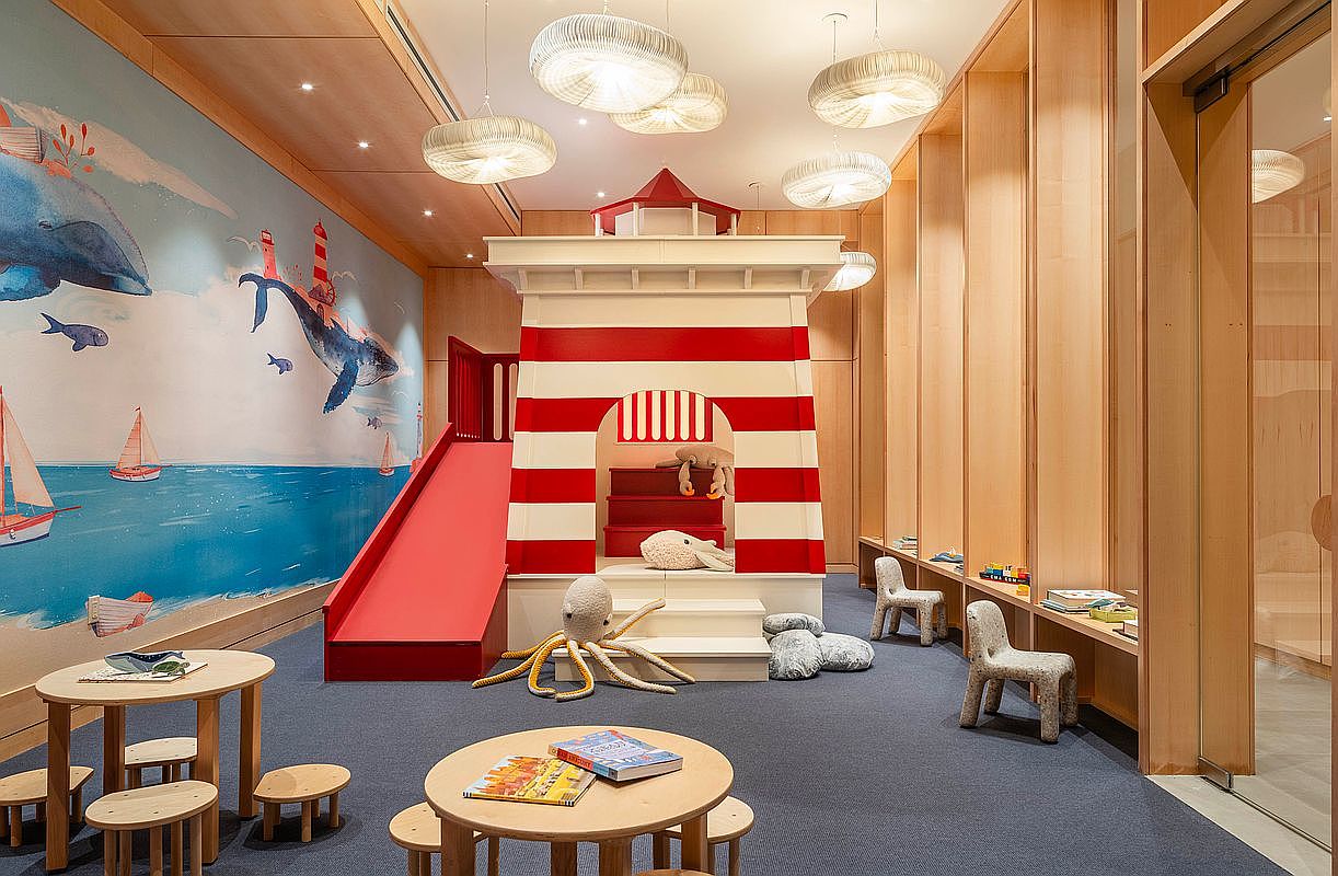 This vibrant children's playroom features a whimsical lighthouse-themed play structure complete with a red slide and nautical-inspired wall murals. The room is furnished with child-sized wooden tables and chairs, soft plush toys, and modern pendant lighting, creating a playful and inviting atmosphere. The space is designed with light wood paneling and a deep blue carpet, emphasizing a clean, high-end aesthetic suitable for a luxury residential amenity space.
