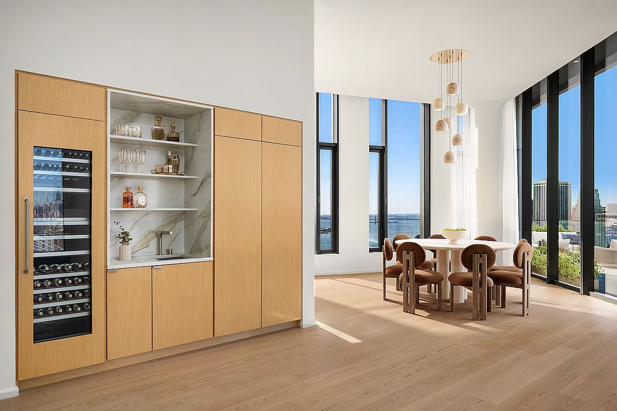 This sophisticated dining area features a modern round table surrounded by plush, velvet-upholstered chairs, illuminated by a cluster of elegant, spherical pendant lights. To the left, a sleek, light-wood built-in unit integrates a wine refrigerator and a marble-backed wet bar, creating a seamless blend of functionality and high-end design. Floor-to-ceiling windows frame expansive city and water views, bathing the room in natural light and emphasizing the open, airy atmosphere of this luxury residence.