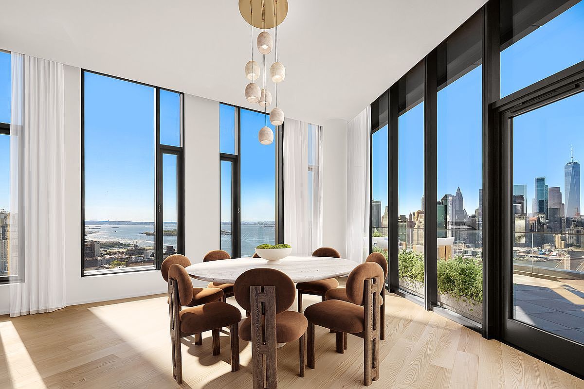 This sophisticated dining room features a round marble-topped table surrounded by modern, velvet-upholstered chairs, set against a backdrop of floor-to-ceiling windows that offer panoramic views of the New York City skyline. A sculptural, multi-pendant light fixture hangs above the table, adding a touch of elegance to the bright, airy space. The room seamlessly blends contemporary interior design with an impressive urban vista, creating a luxurious and inviting atmosphere.