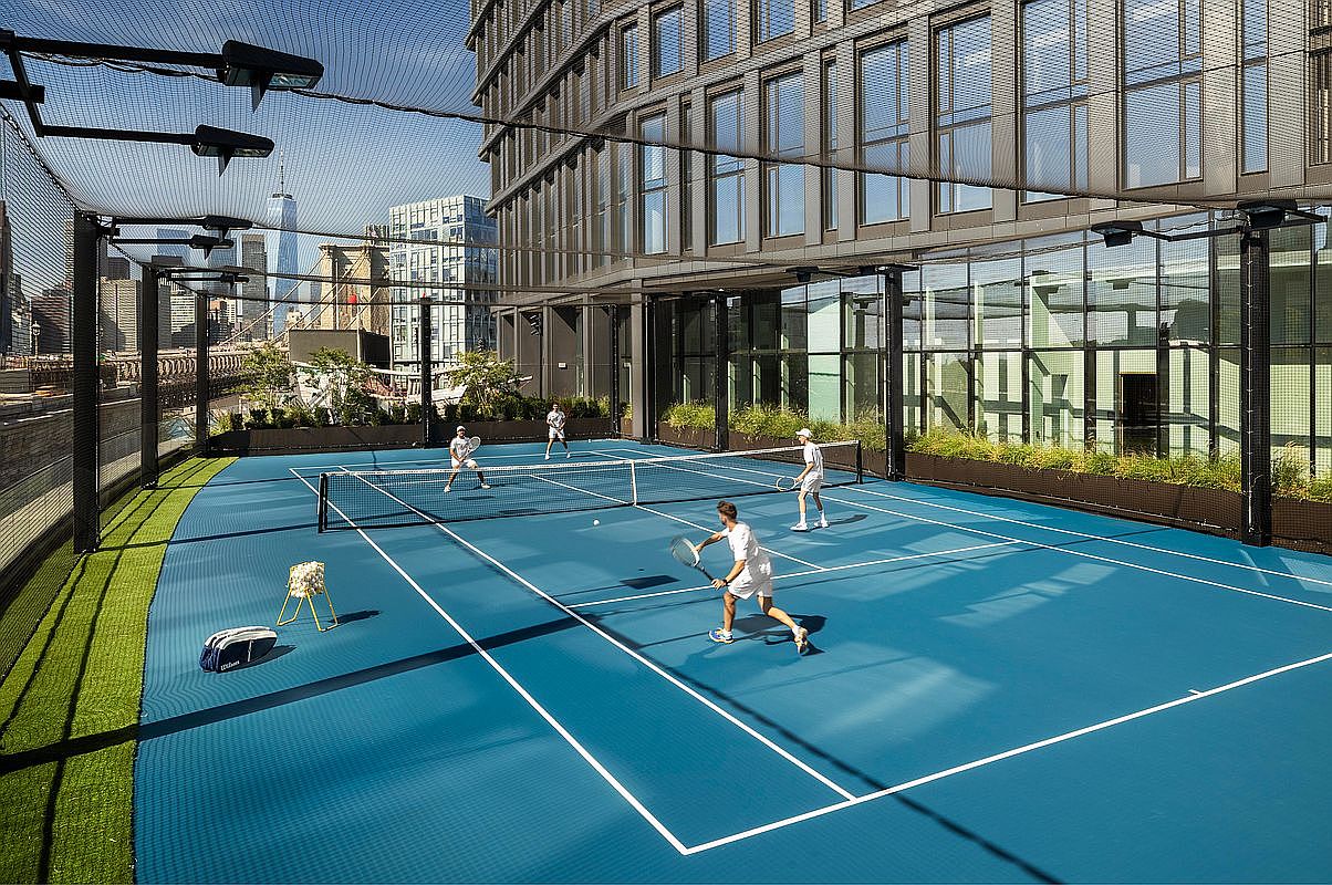 This image showcases a high-end, rooftop tennis court featuring a vibrant blue surface and professional netting, set against the backdrop of a modern glass-clad building. The perspective captures players in action, emphasizing the luxury lifestyle and recreational amenities available to residents. The urban setting is highlighted by the iconic city skyline visible in the distance, creating an impressive and desirable atmosphere.