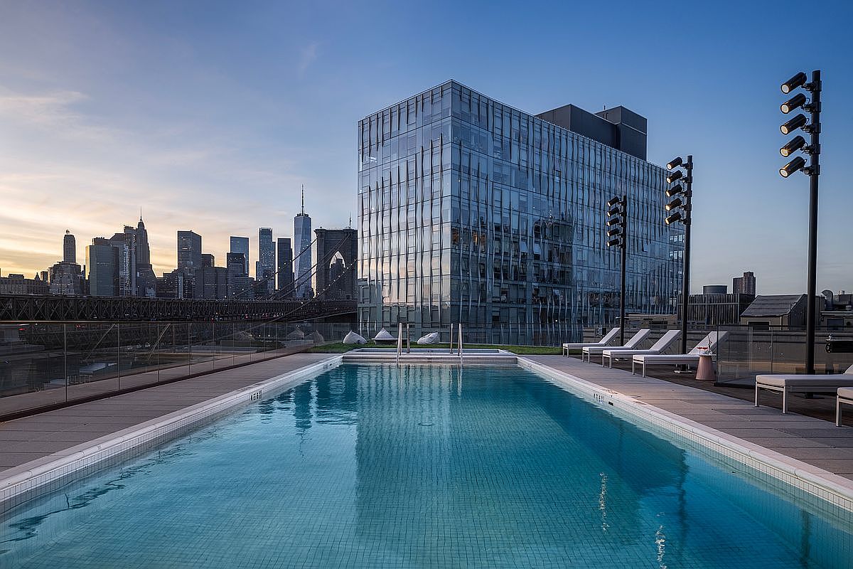 This stunning rooftop pool offers a breathtaking panoramic view of the New York City skyline, including the iconic Brooklyn Bridge. The sleek, modern deck is furnished with minimalist lounge chairs and tall, contemporary lighting fixtures, creating a sophisticated urban oasis. The composition captures the serene blue water of the pool in the foreground, contrasting beautifully with the glass-clad architecture and the vibrant city backdrop at dusk.