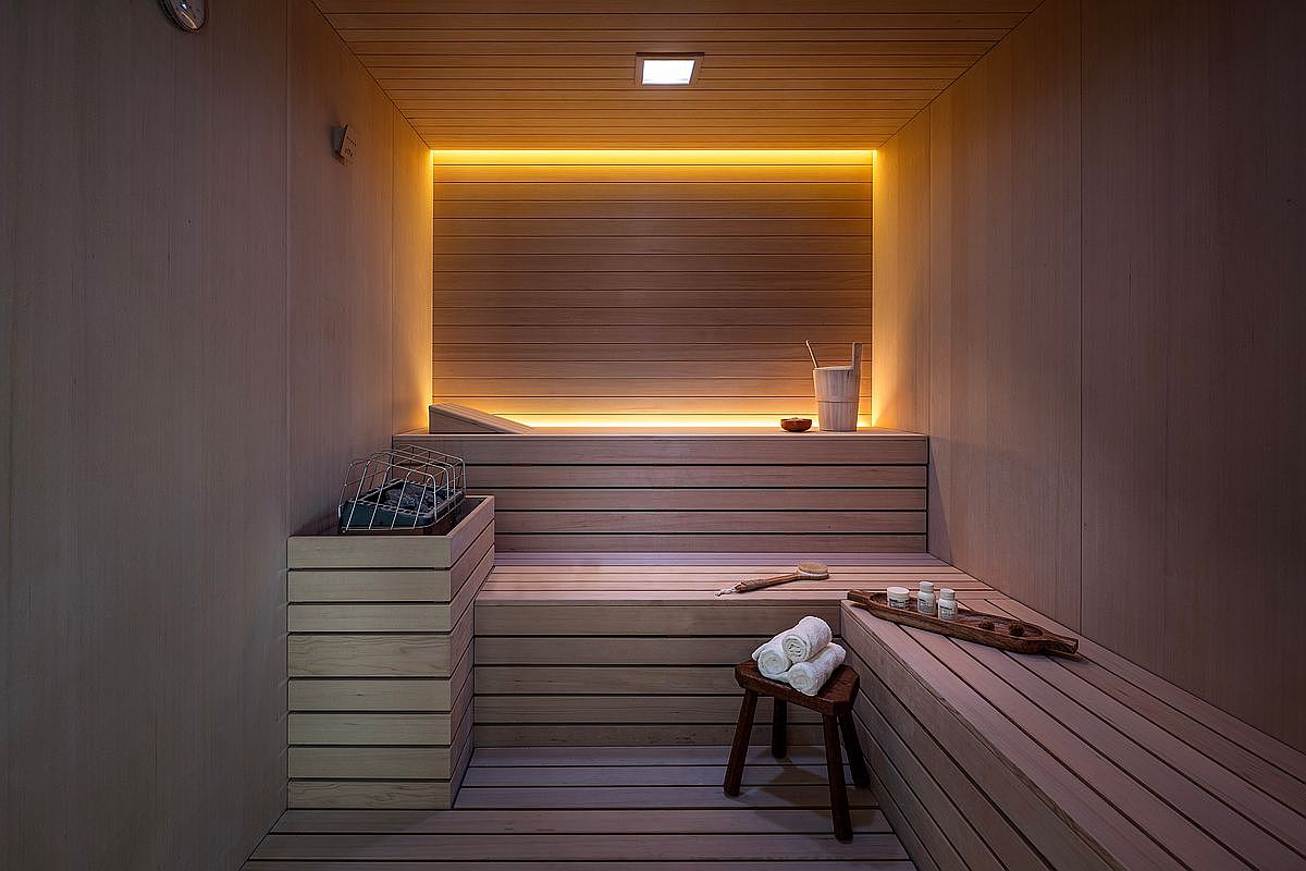 This image showcases a minimalist, modern sauna interior characterized by warm wood paneling and soft, recessed ambient lighting. The space features tiered wooden benches, a dedicated heater unit, and a small wooden stool with rolled towels, creating a serene and luxurious spa-like atmosphere. The perspective is a centered, eye-level shot that emphasizes the clean lines and tranquil aesthetic of this wellness amenity.