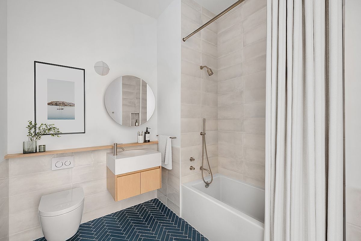 This modern, compact bathroom features a striking navy blue herringbone tile floor that contrasts beautifully with the light, neutral-toned wall tiles. The space includes a floating light wood vanity, a circular mirror, and a combined shower-tub area with a sleek curtain. The minimalist design is accented by a floating shelf and a wall-mounted toilet, creating a clean and contemporary aesthetic.