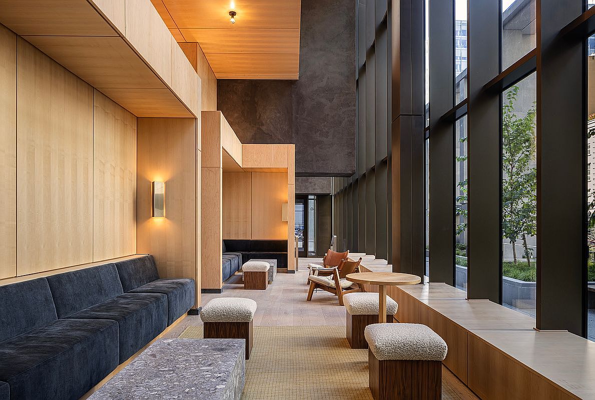 This sophisticated lounge area features a blend of warm wood paneling, dark velvet seating, and textured stone accents, creating a high-end, modern atmosphere. Large floor-to-ceiling windows provide an expansive view of an outdoor garden, while the open layout includes various seating arrangements like plush ottomans and mid-century modern chairs. The space is designed for comfort and social interaction, offering a refined aesthetic suitable for a luxury residential building's common area.