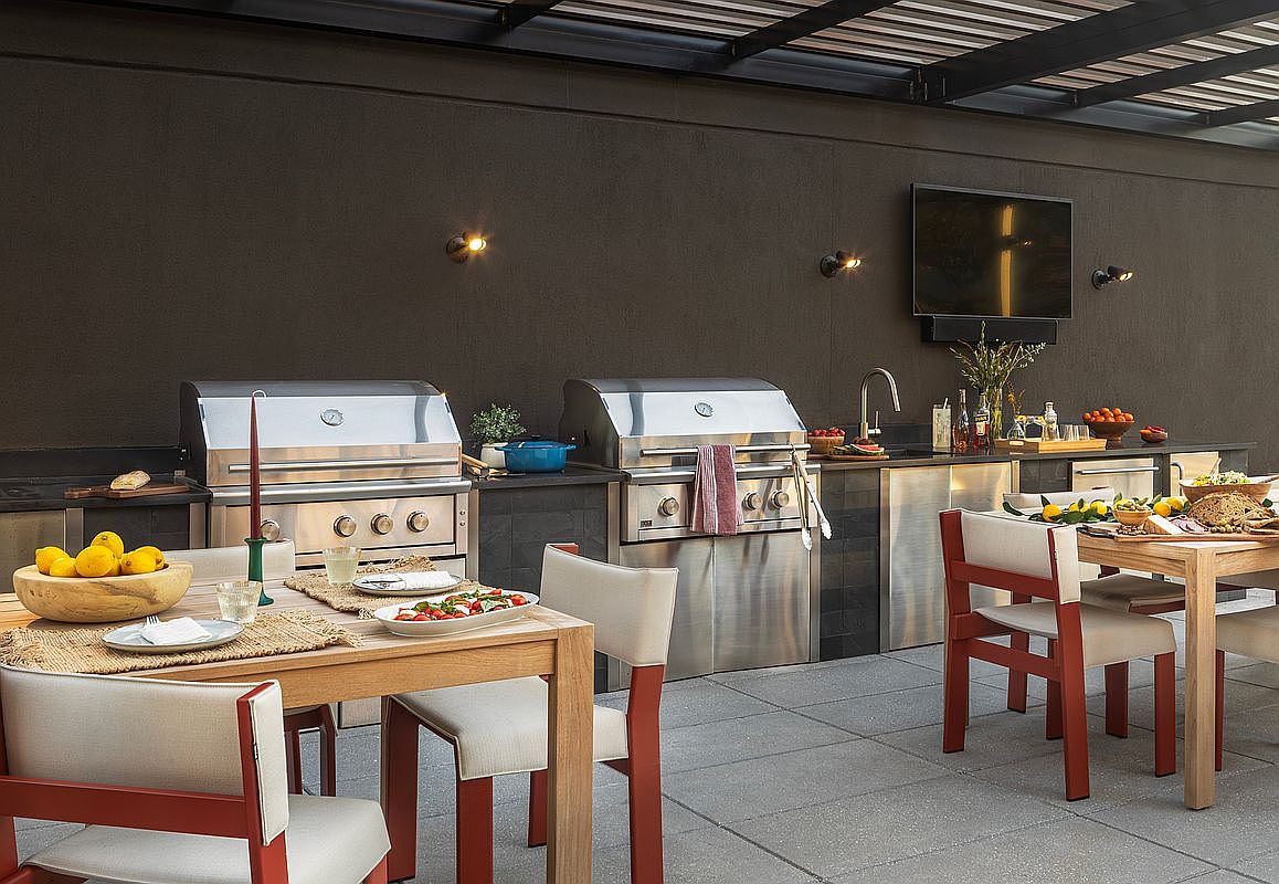 This sophisticated outdoor kitchen and dining area features dual stainless steel grills, sleek dark stone countertops, and a mounted television, perfect for high-end entertaining. The space is furnished with two wooden dining sets and modern chairs, all set against a dark accent wall under a covered patio structure. The overall aesthetic is contemporary and luxurious, emphasizing a seamless transition between indoor comfort and outdoor living.