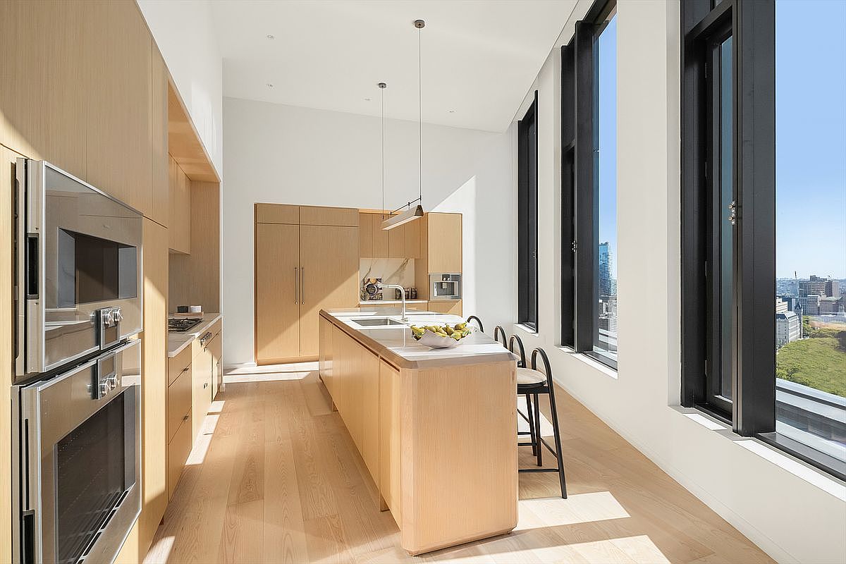 This sleek, modern kitchen features light wood cabinetry, a central island with a stone countertop, and high-end stainless steel appliances. The space is bathed in natural light from floor-to-ceiling windows that offer a stunning, expansive view of the city skyline and greenery below. The minimalist design and high ceilings create an airy, sophisticated atmosphere perfect for contemporary urban living.
