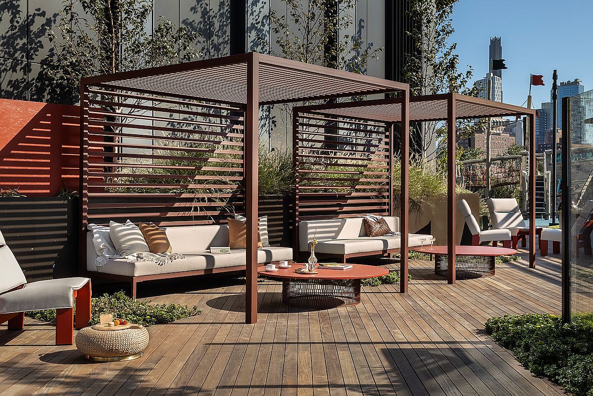 This sophisticated rooftop terrace features two modern, slatted-wood cabanas housing comfortable outdoor lounge seating and circular coffee tables. The space is finished with high-quality wood decking, lush greenery, and a clear view of the urban skyline, creating an upscale, tranquil retreat. The cinematic perspective captures the inviting, sun-drenched atmosphere, perfect for luxury outdoor entertaining.