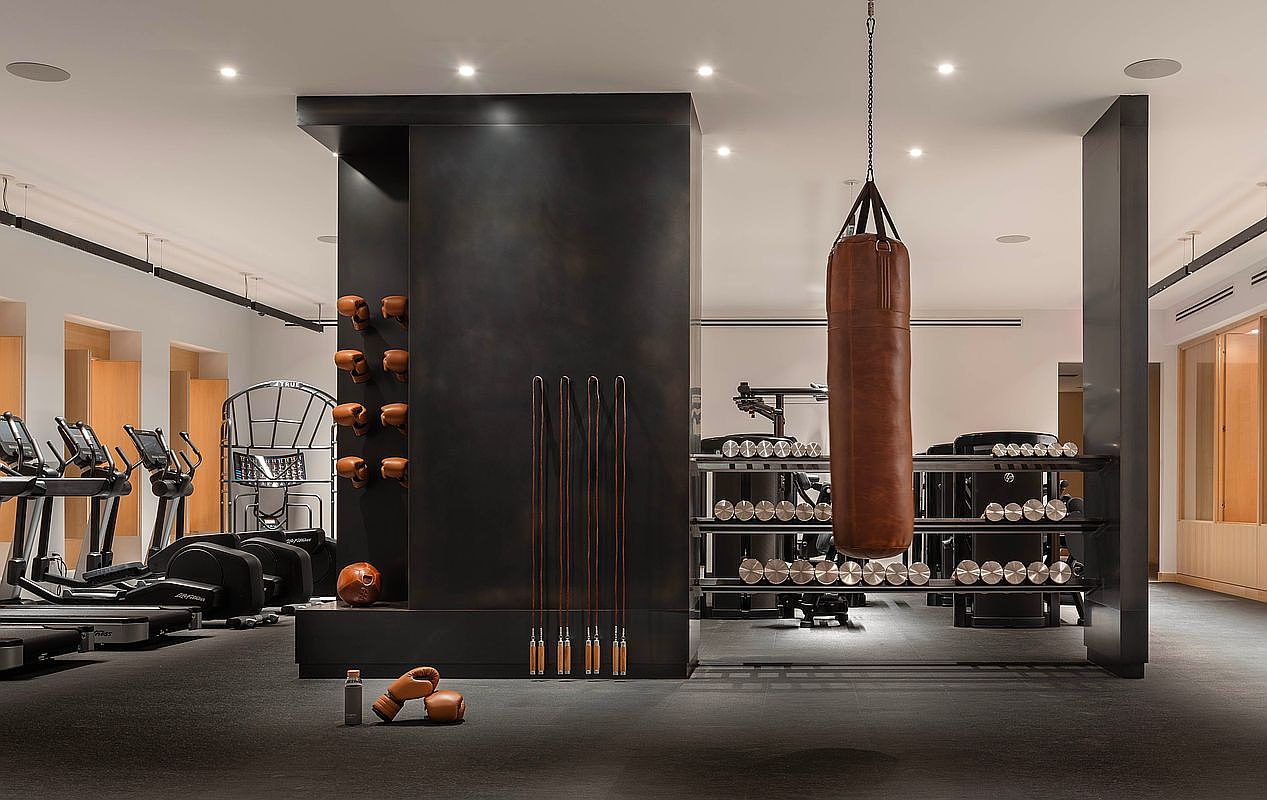 This sophisticated, high-end home gym features a sleek, dark architectural pillar as its centerpiece, adorned with leather boxing gloves and jump ropes. The space is equipped with professional-grade cardio machines, a heavy leather punching bag, and organized racks of dumbbells, all set against a minimalist, modern aesthetic. The cinematic perspective captures the gym's organized layout and premium finishes, conveying an atmosphere of luxury and focused fitness.