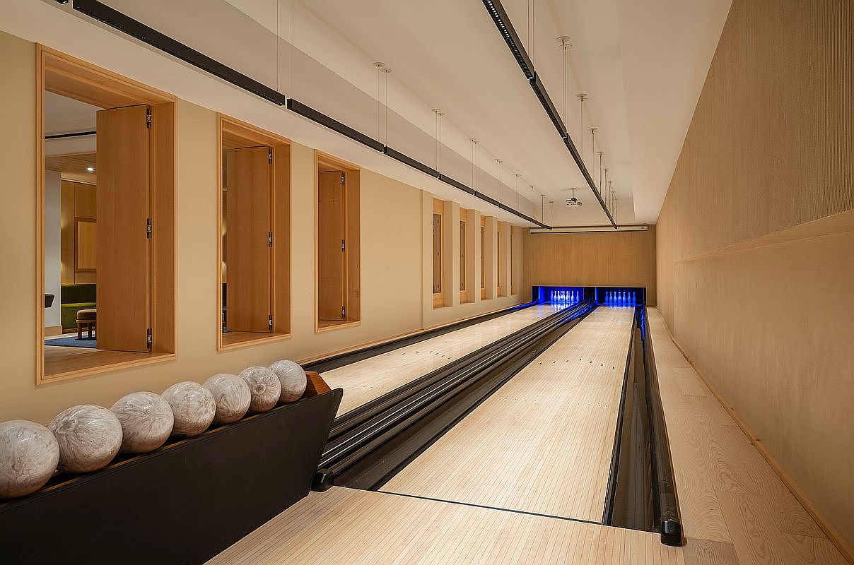 This image showcases a sleek, modern private bowling alley featuring two lanes with polished wooden surfaces and glowing blue pin-setting areas. The room is designed with minimalist light wood paneling, recessed lighting, and a row of bowling balls resting on a rack in the foreground. The perspective is a wide-angle shot looking down the length of the lanes, emphasizing the clean lines and high-end recreational atmosphere.