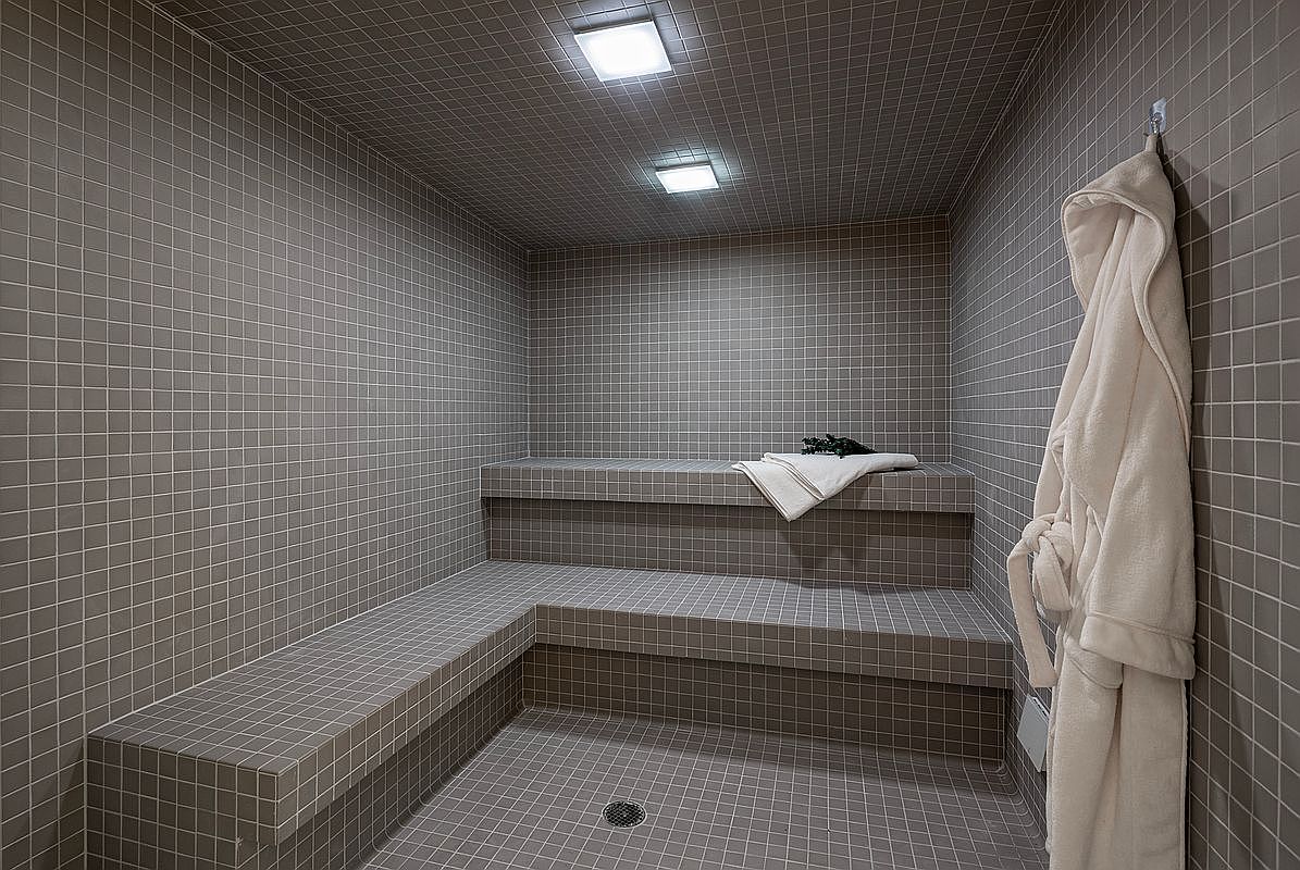 This image showcases a sleek, modern steam room or sauna completely tiled in a neutral, grid-patterned gray ceramic. The space features built-in tiered benches, a floor drain, and a plush white bathrobe hanging on the wall, creating a spa-like atmosphere. The perspective is a wide-angle interior shot that emphasizes the clean lines and minimalist design of this luxury wellness amenity.