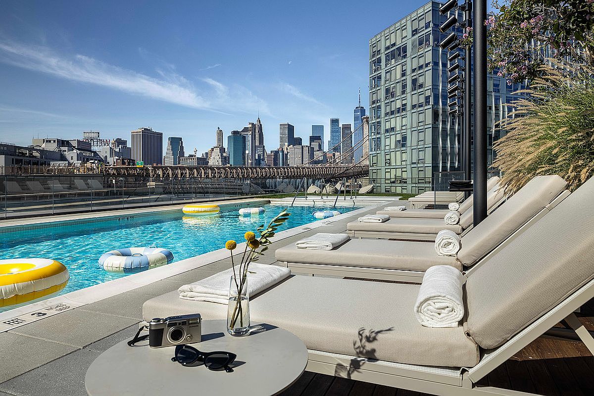 This stunning rooftop pool area offers a luxurious urban retreat with a direct, iconic view of the New York City skyline and the Brooklyn Bridge. The space is furnished with sleek, modern lounge chairs, white towels, and a small side table holding a camera and sunglasses, creating a sophisticated and relaxing atmosphere. The bright blue water of the pool contrasts beautifully with the architectural backdrop, making it an ideal setting for high-end city living.