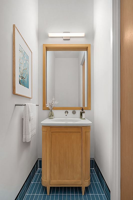 This compact powder room features a warm, light-wood vanity with a clean white countertop and a matching framed mirror. The space is accented by striking deep blue floor tiles and a minimalist wall-mounted light fixture, creating a modern and sophisticated aesthetic. A framed piece of art on the wall and a neatly hung towel add a touch of refined, functional style to this small bathroom.