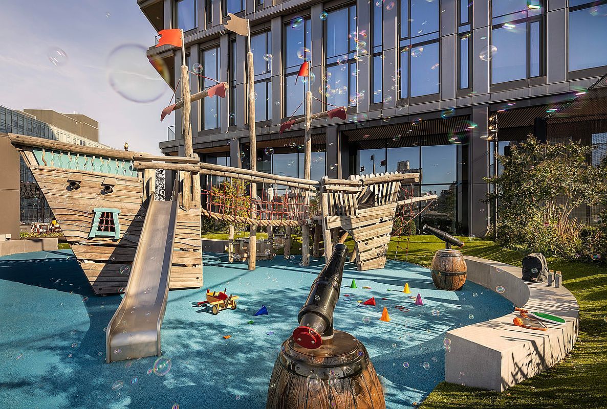 This vibrant outdoor playground features a large, custom-built wooden pirate ship structure complete with a slide, climbing nets, and decorative cannons. Set against a modern, multi-story glass-facade building, the area is filled with playful bubbles, creating a whimsical and family-friendly atmosphere. The ground is covered in a soft, blue safety surface, making it an ideal recreational space for children within a residential community.