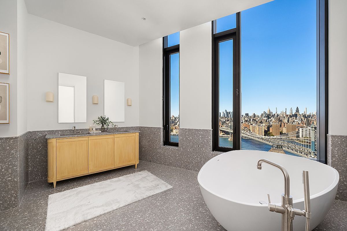 This luxurious primary bathroom features a sleek, modern design with a freestanding soaking tub positioned to capture a breathtaking panoramic view of the city skyline and bridge. The space is anchored by a light wood double vanity with stone countertops and elegant wall-mounted mirrors, all set against sophisticated grey terrazzo-style flooring and wall tiling. The floor-to-ceiling windows flood the room with natural light, creating an airy and high-end atmosphere.