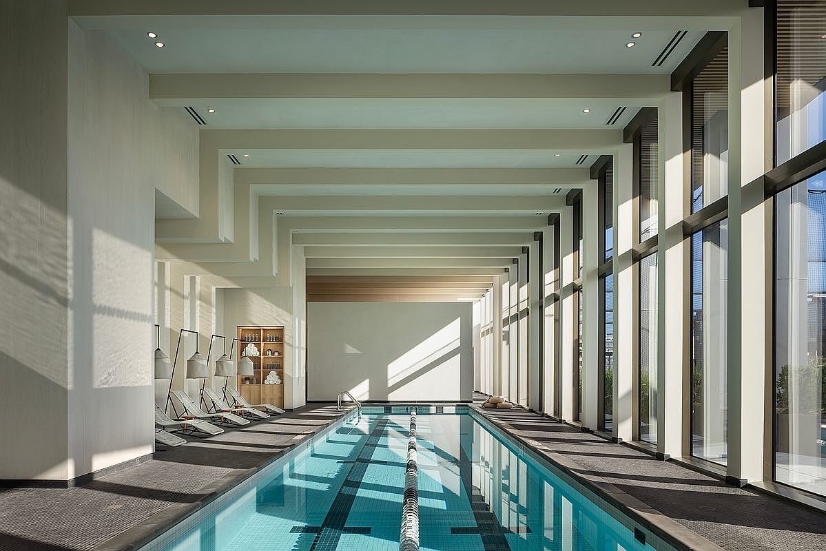 This sophisticated indoor lap pool area features a sleek, minimalist design with high ceilings, exposed structural beams, and floor-to-ceiling windows that bathe the space in natural light. The room includes a row of modern lounge chairs along the side, creating a serene and luxurious atmosphere perfect for relaxation or exercise. The perspective is a wide, symmetrical shot looking down the length of the pool, emphasizing the clean lines and architectural elegance of the facility.