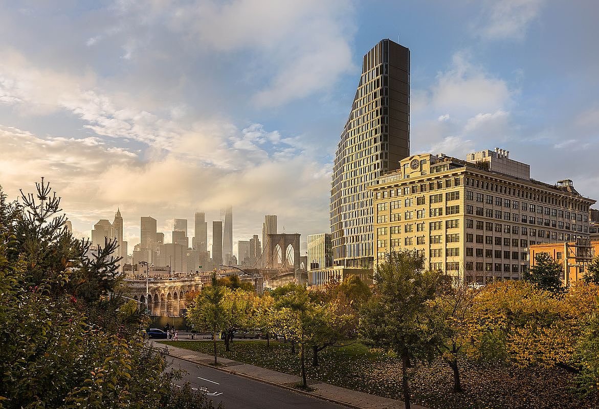 This cinematic wide shot captures a vibrant urban park in the foreground, transitioning into a striking architectural contrast between a historic masonry building and a modern, angular glass skyscraper. In the distance, the iconic Brooklyn Bridge and the hazy Manhattan skyline create a dramatic, high-value backdrop. The scene is bathed in the warm, golden light of late afternoon, emphasizing the blend of nature and metropolitan density.