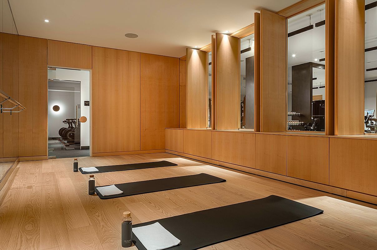 This serene, minimalist yoga or fitness studio features warm wood-paneled walls and matching light-toned hardwood flooring. Three black yoga mats are neatly laid out on the floor, each accompanied by a water bottle and a folded white towel, creating an inviting and organized atmosphere. The room is illuminated by recessed lighting and features large glass partitions that offer a glimpse into an adjacent gym area, enhancing the sense of depth and modern luxury.