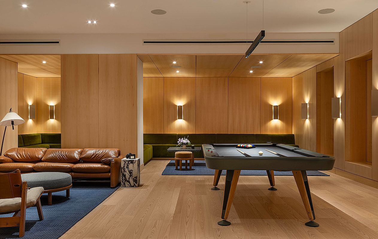 This sophisticated lounge area features a modern pool table as the centerpiece, complemented by a plush leather sofa and integrated wood-paneled walls. The space is illuminated by warm, ambient wall sconces and recessed lighting, creating an inviting atmosphere for social gatherings. The clean lines, natural wood tones, and minimalist aesthetic suggest a high-end residential amenity space or clubhouse.