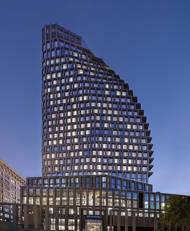 This striking high-rise building features a unique, stepped architectural design with a dark, grid-like facade that creates a modern and sophisticated aesthetic. The structure is captured from a low-angle perspective during twilight, highlighting the warm, inviting glow of the interior lights against the deep blue sky. The building's tiered silhouette and expansive glass windows suggest a luxury residential or commercial development with a contemporary urban appeal.