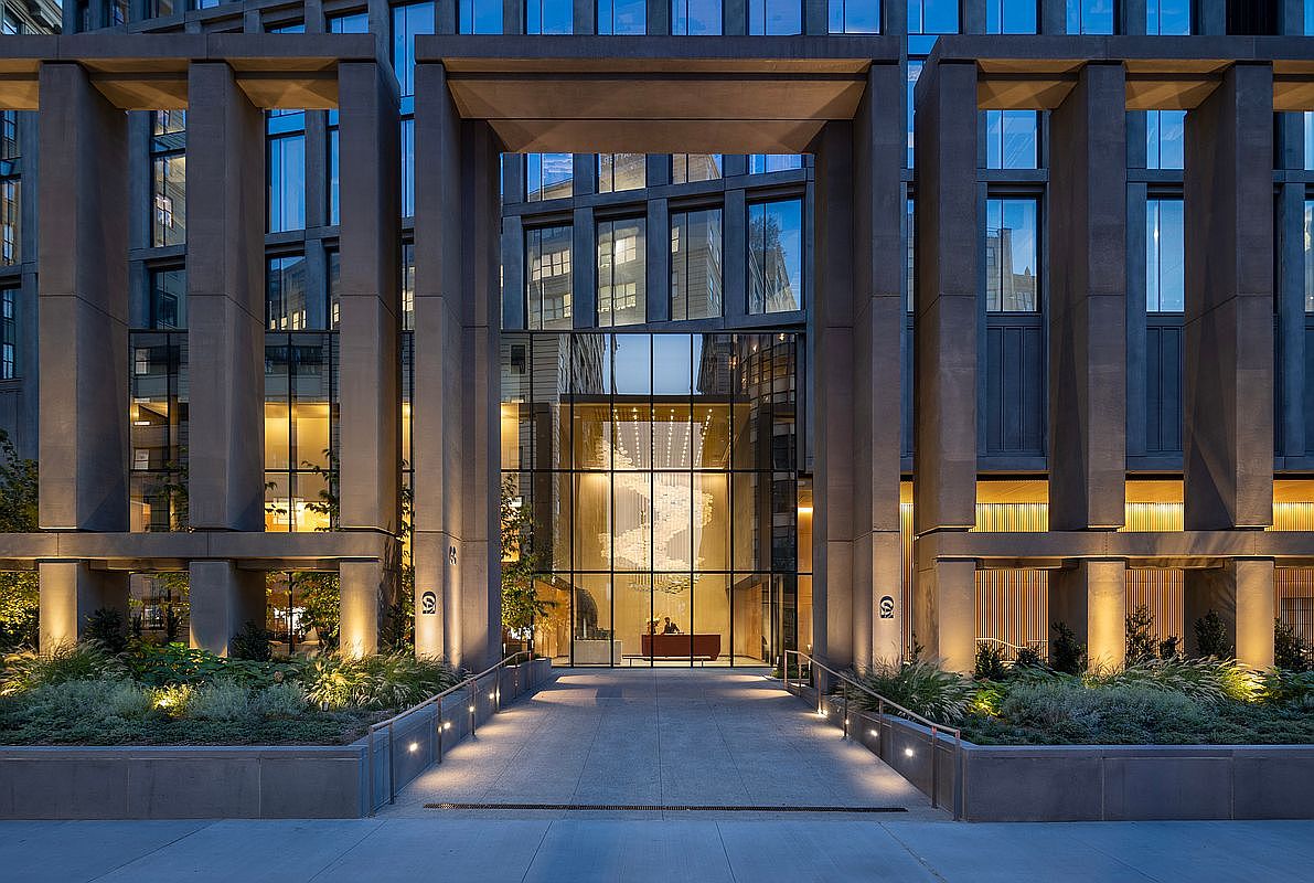 This striking, modern entryway features a grand, symmetrical stone facade with towering columns and a central glass-enclosed lobby. A paved walkway leads directly to the entrance, flanked by manicured landscaping and warm, integrated ground lighting that highlights the building's architectural texture. The perspective is centered and cinematic, emphasizing the imposing scale and sophisticated, high-end urban aesthetic of the property.