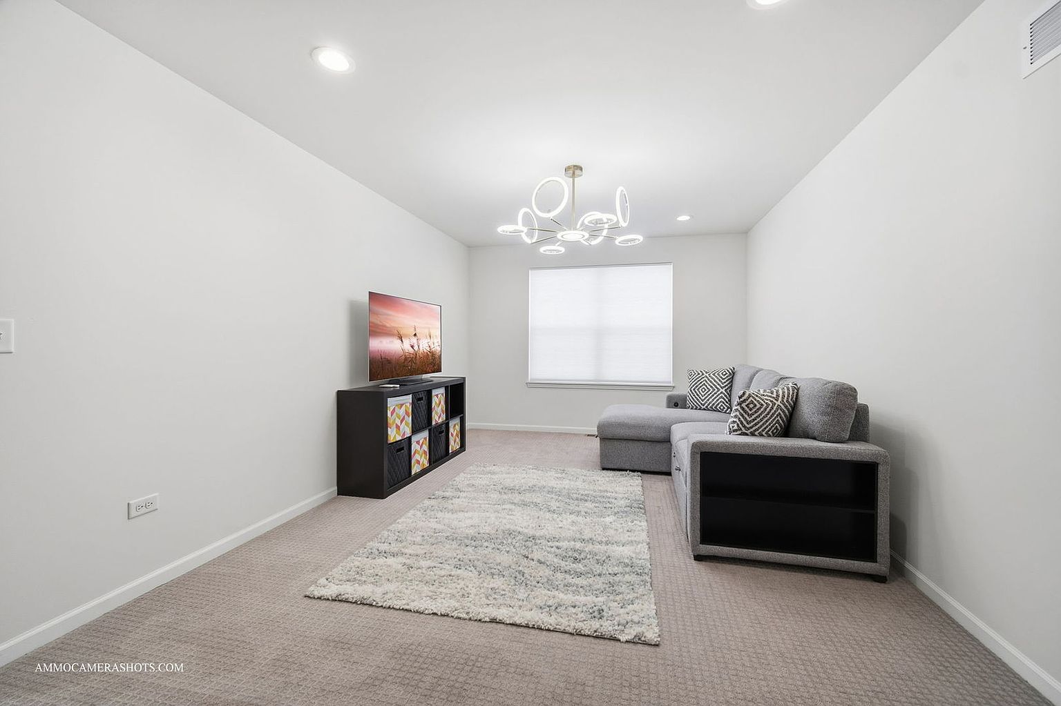 This is a bright and modern living room featuring a gray sectional sofa with decorative pillows, a contemporary chandelier, and a flat-screen TV on a black media console. A patterned area rug adds texture to the neutral carpet, and a large window provides natural light. The room has a clean and minimalist aesthetic, creating a comfortable and inviting space.