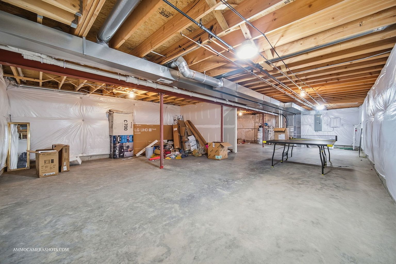 This is a spacious unfinished basement with a concrete floor and exposed wooden ceiling beams. The walls are partially covered with white plastic sheeting, and there are several cardboard boxes and a ping pong table in the room. The space appears to be well-lit and offers potential for customization and finishing.