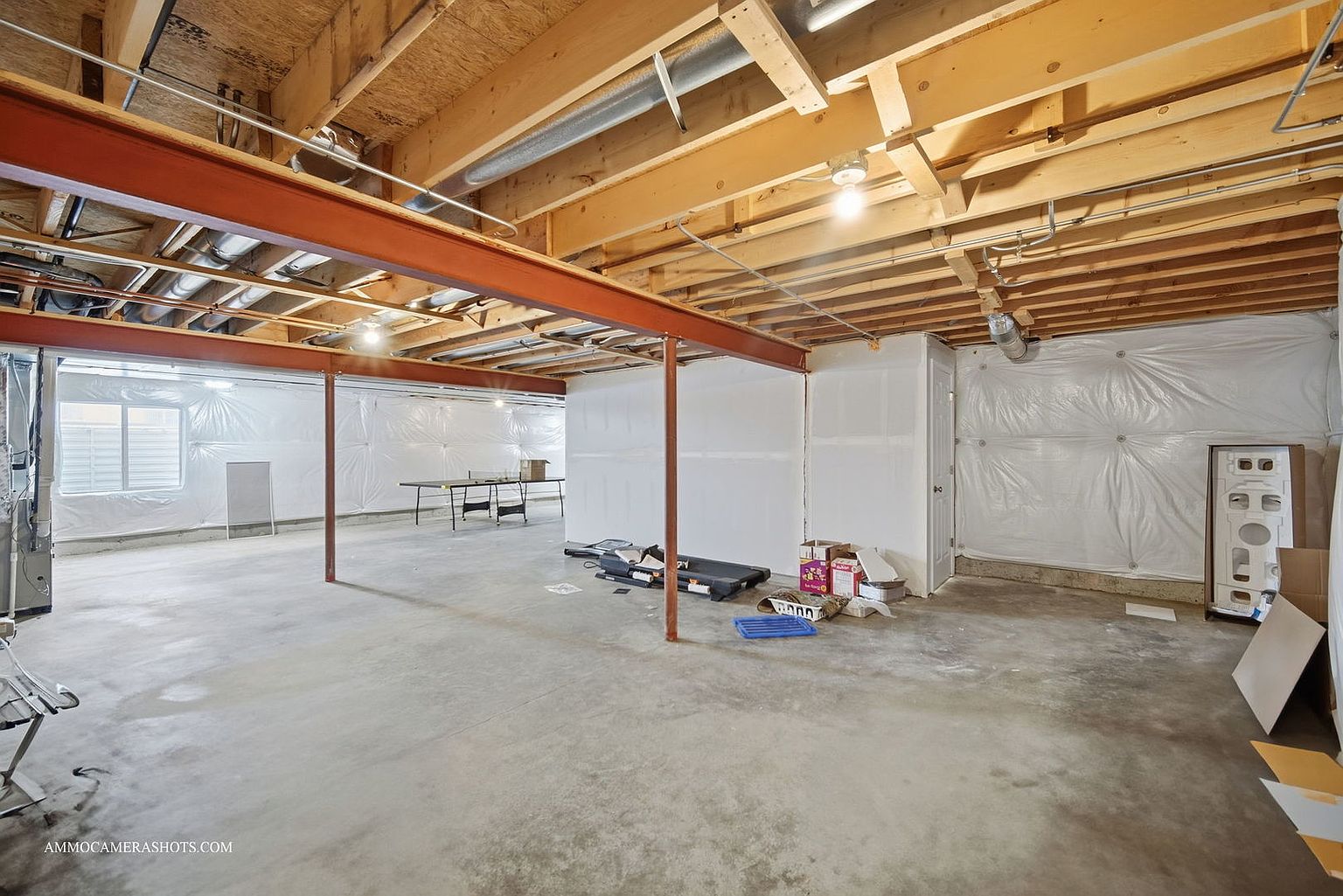 This is an unfinished basement with exposed wooden ceiling beams and a concrete floor. The walls are partially covered with white plastic sheeting, and a large steel beam runs across the ceiling, supported by metal posts. There are some boxes and equipment scattered around, suggesting a space ready for customization.