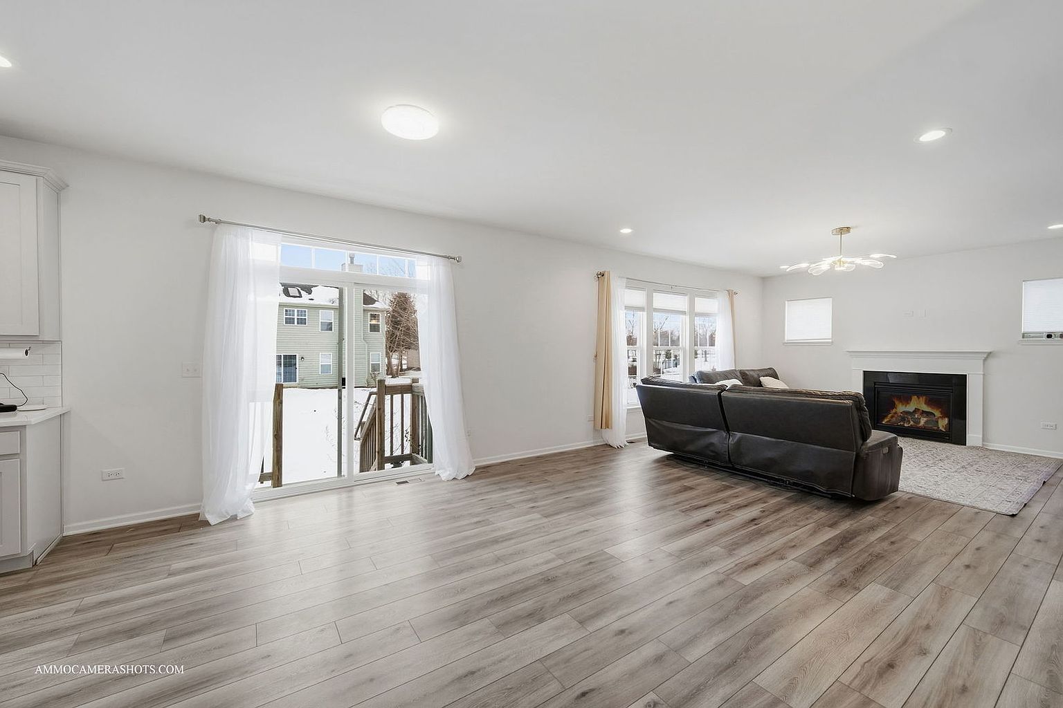 This is a bright and airy living room featuring light wood flooring and white walls. A dark leather sofa sits in the center of the room, facing a fireplace with a white mantel. Large windows and a sliding glass door provide ample natural light, enhancing the spacious feel of the room.