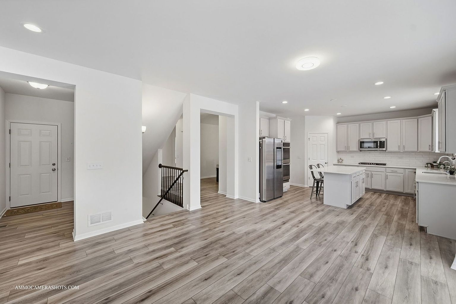 This is an interior shot of a modern, open-concept kitchen and living area. The kitchen features white cabinets, stainless steel appliances, and a central island. The flooring is a light-colored wood laminate, and the walls are painted a neutral white, creating a bright and airy atmosphere. The perspective is from the living area, showcasing the seamless flow between the kitchen and the rest of the home.