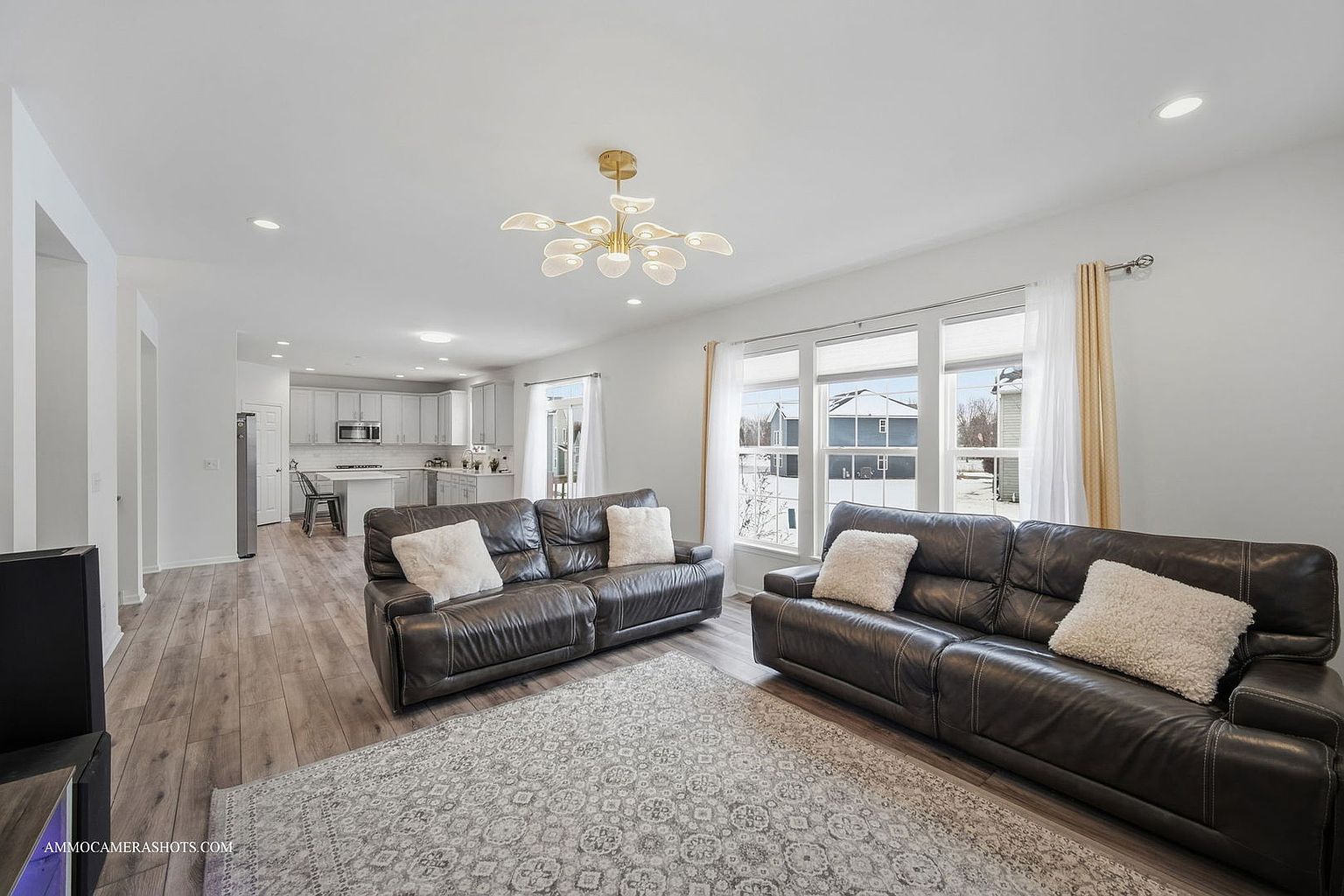 This is a bright and spacious living room featuring two dark leather sofas with light-colored throw pillows, a patterned area rug, and large windows that provide ample natural light. The open floor plan seamlessly connects to the kitchen, creating a welcoming and modern living space. A stylish chandelier hangs from the ceiling, adding a touch of elegance.