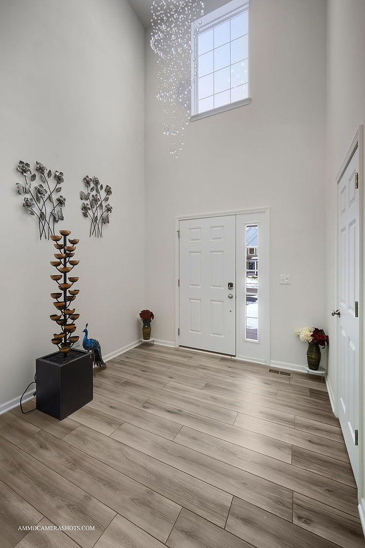 This is an interior shot of a bright and spacious entryway. The walls are painted in a neutral tone, complemented by light wood-look flooring. A decorative water fountain and metal floral wall art add visual interest, while a large window above the front door allows ample natural light to flood the space.
