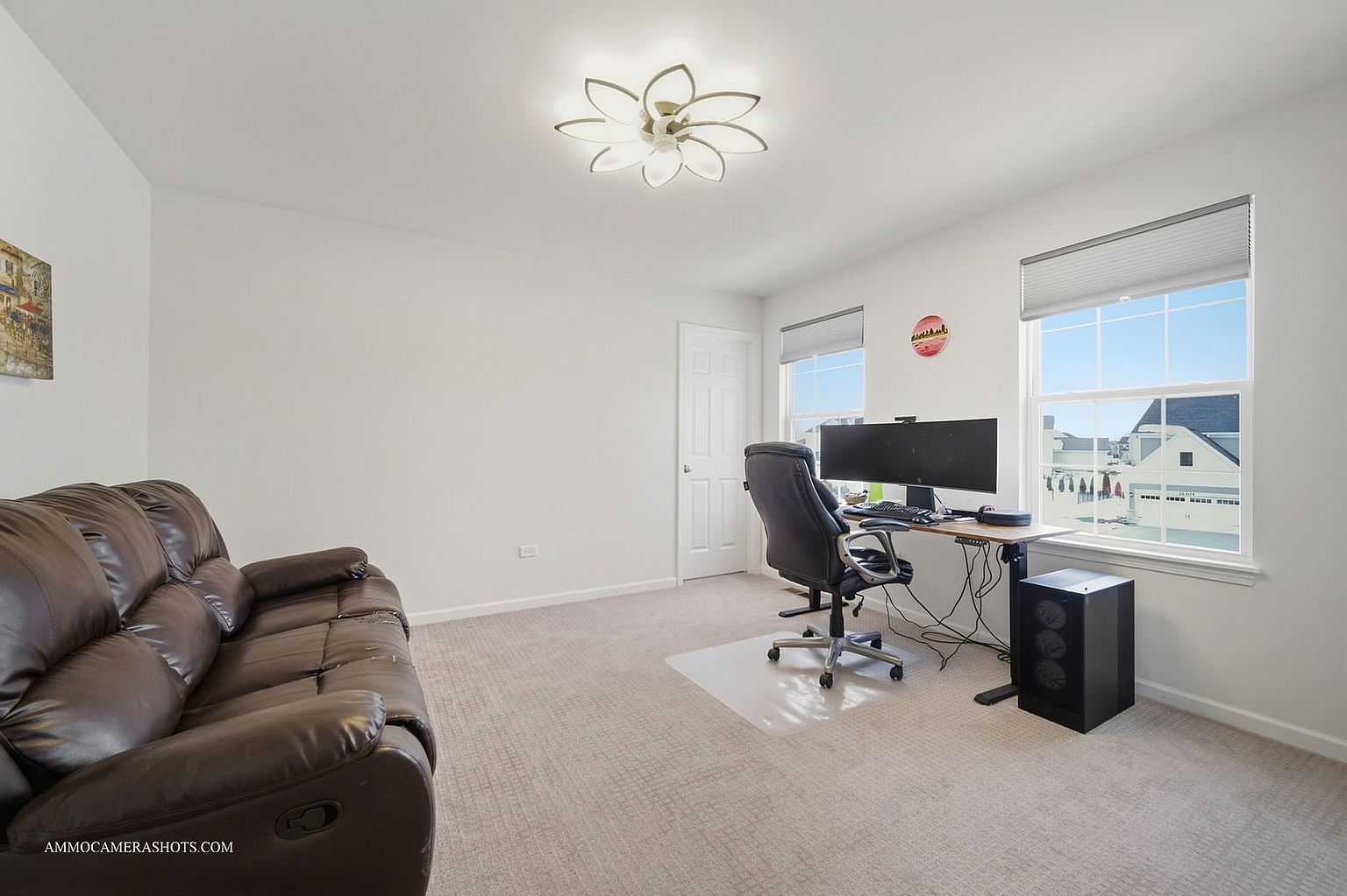 This is a bright and tidy home office featuring a brown leather sofa, a computer desk setup with a large monitor, and a modern ceiling light fixture. The room has neutral-colored carpet and white walls, creating a clean and functional workspace. Natural light streams in through the windows, enhancing the airy feel of the room.