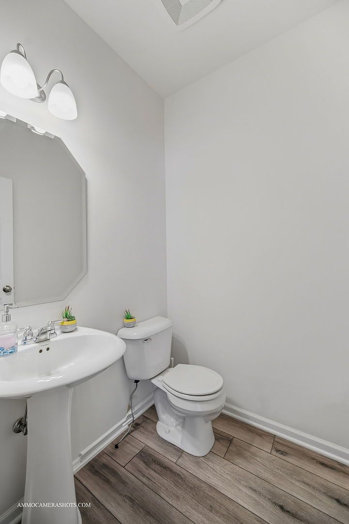 This is a well-lit, clean guest bathroom featuring a pedestal sink with a chrome faucet, an octagonal mirror, and a standard toilet. The walls are painted in a neutral tone, and the flooring is wood-look laminate. The overall impression is simple and functional.