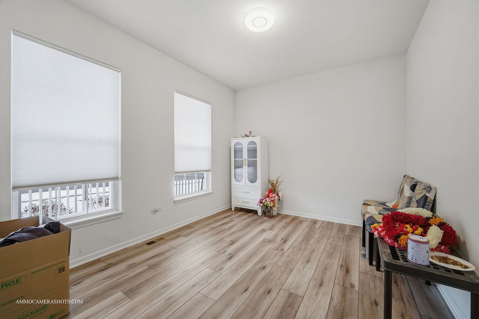 The image showcases a bright, sparsely furnished room with light wood-look flooring and white walls. Two windows with white blinds provide natural light. A white cabinet stands against one wall, and a small table with floral arrangements and other items sits near a chair in the corner, creating a clean and versatile space.